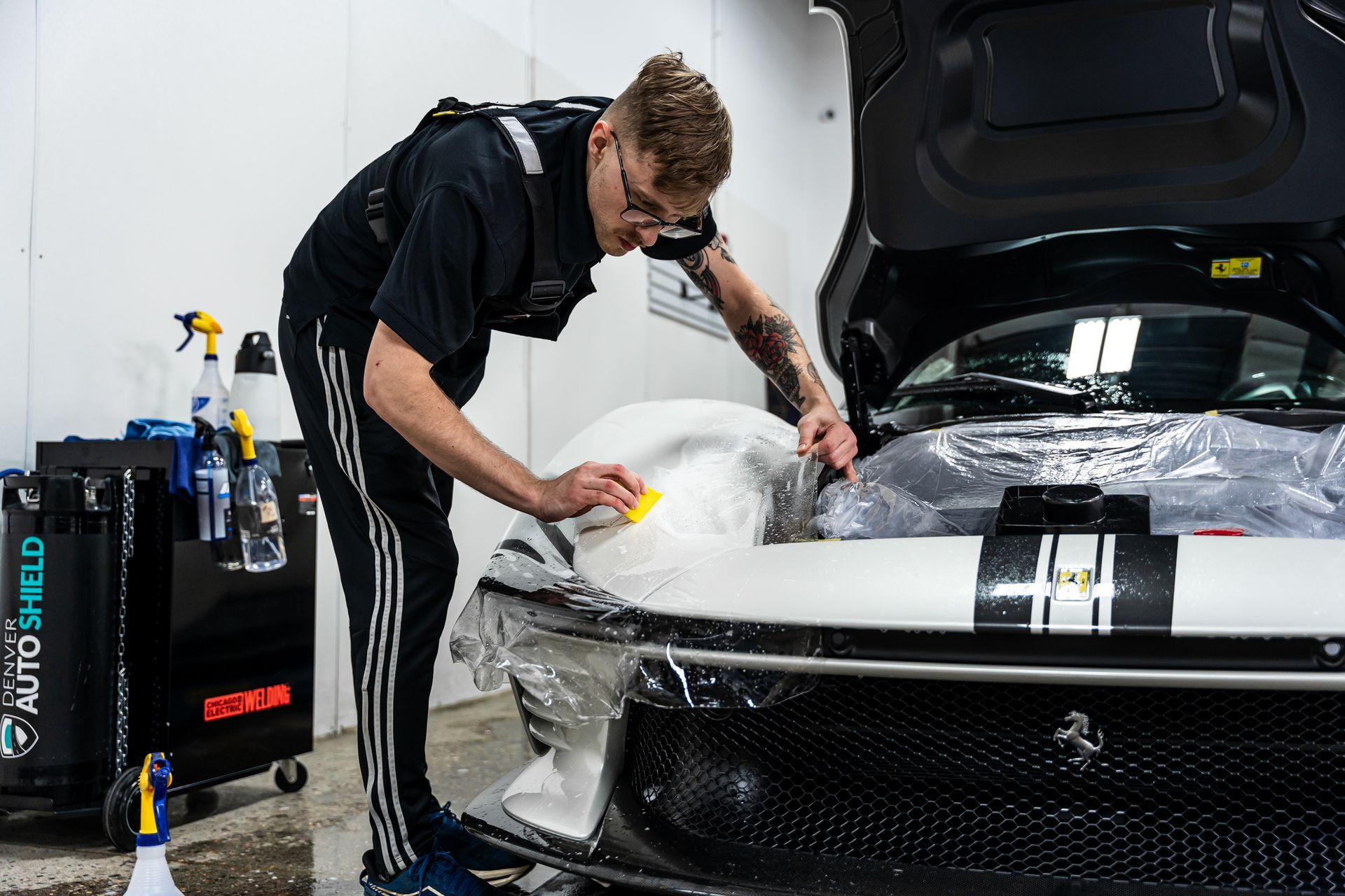 Man applying protective film to the hood of a black and white sports car in a garage.