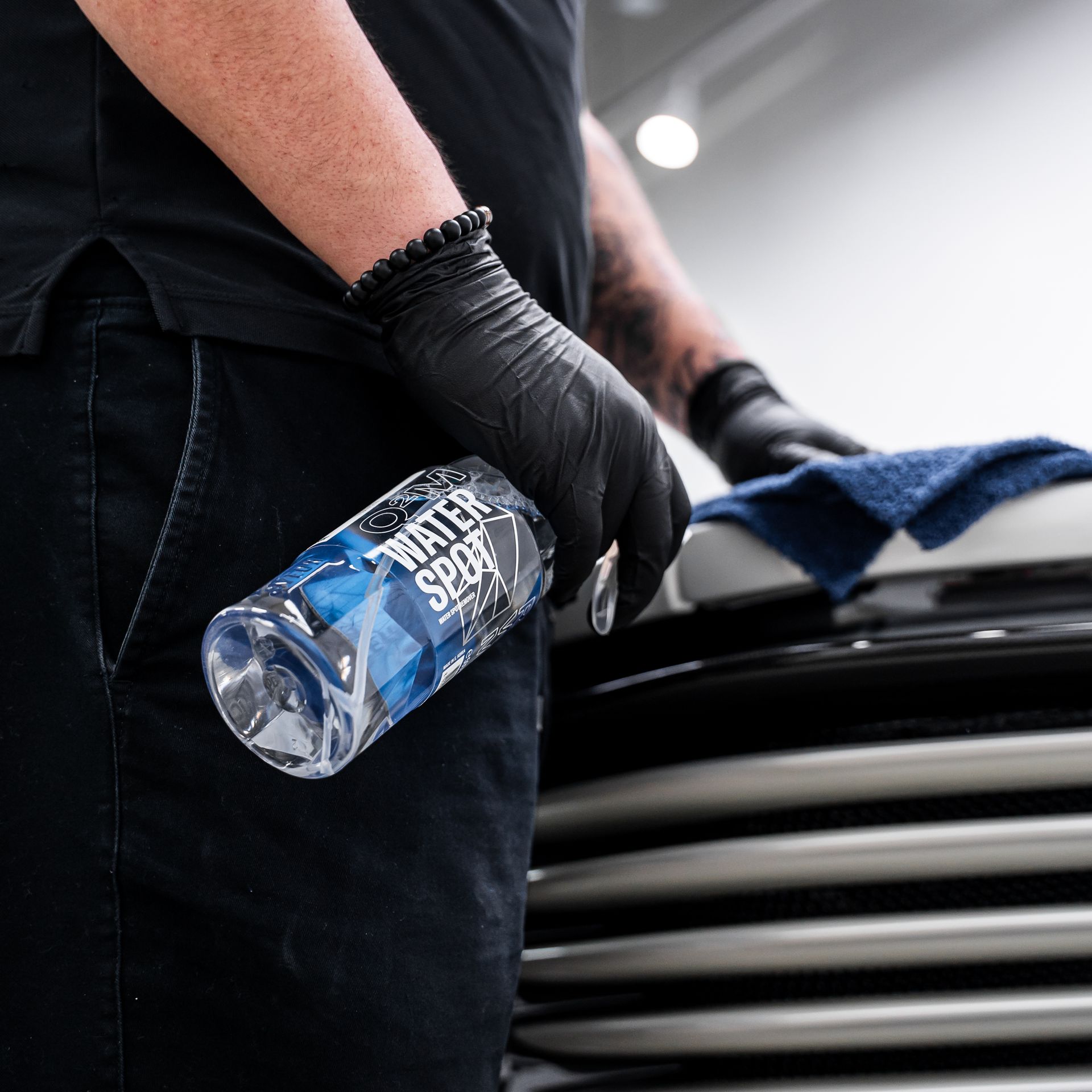 Person in black gloves, holding a water spot remover, wiping a car's grill with a blue cloth.