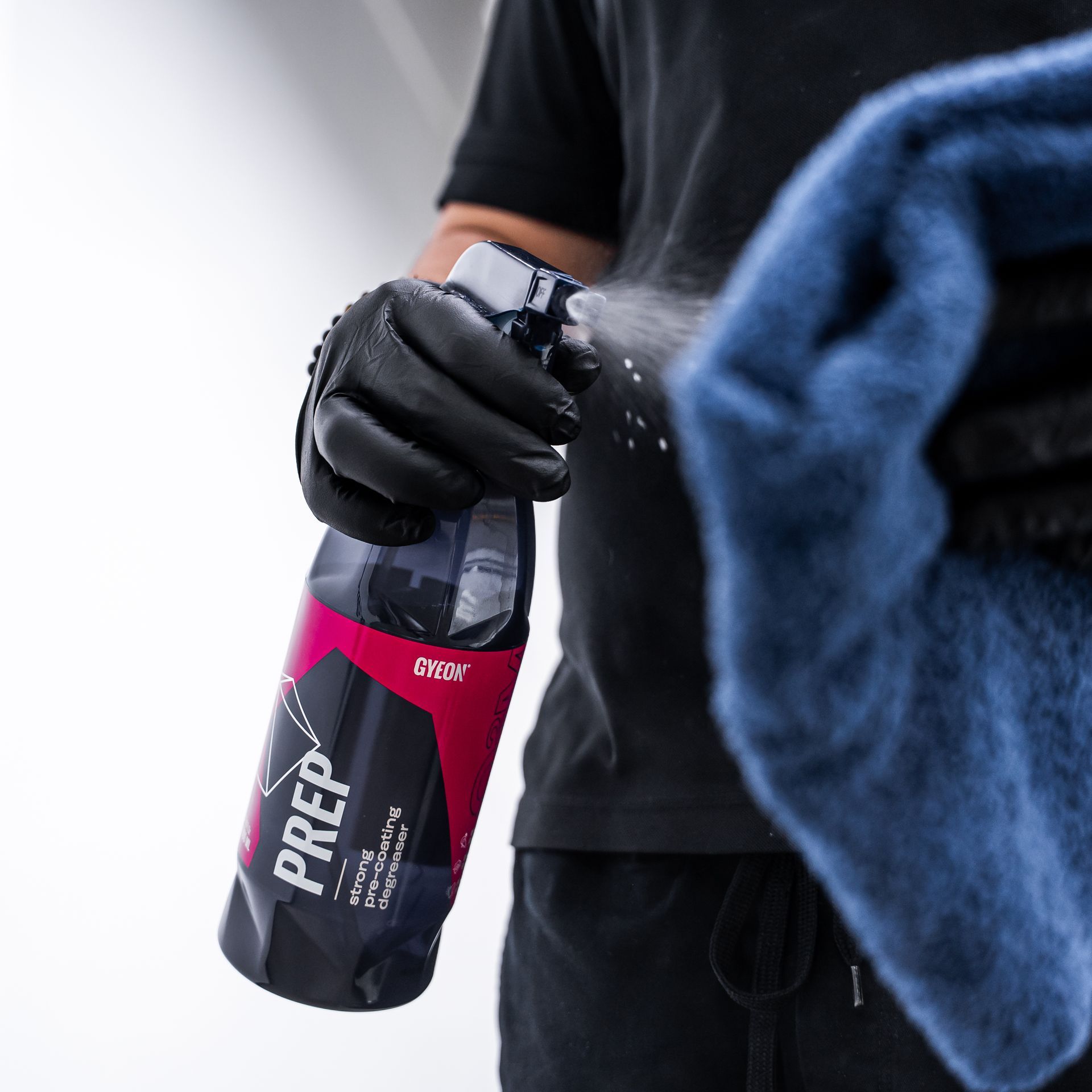 Person spraying car prep product onto a blue microfiber towel while wearing black gloves and a black shirt.