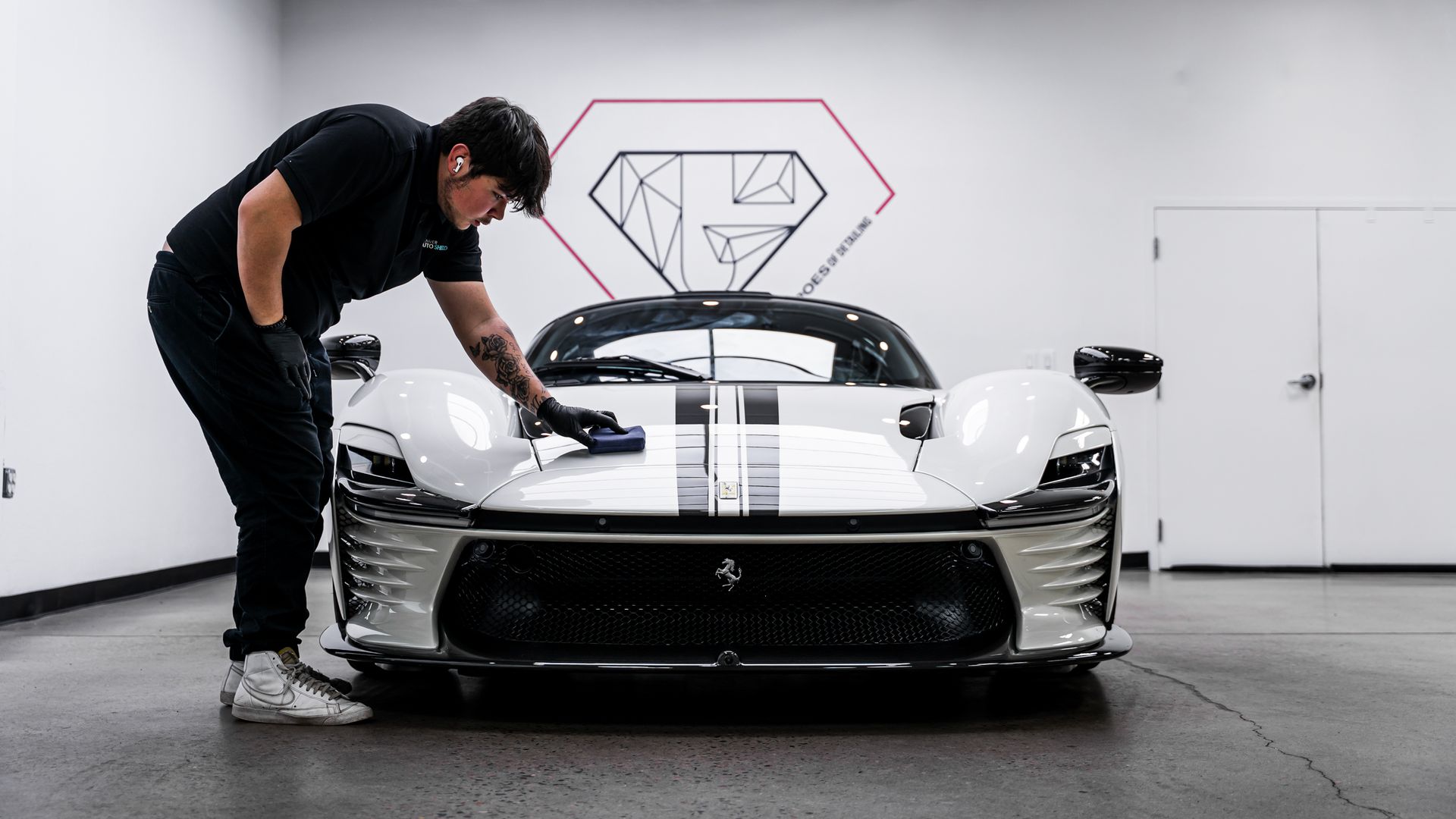 Man detailing a white sports car with black stripes, in a garage with a logo on the wall.