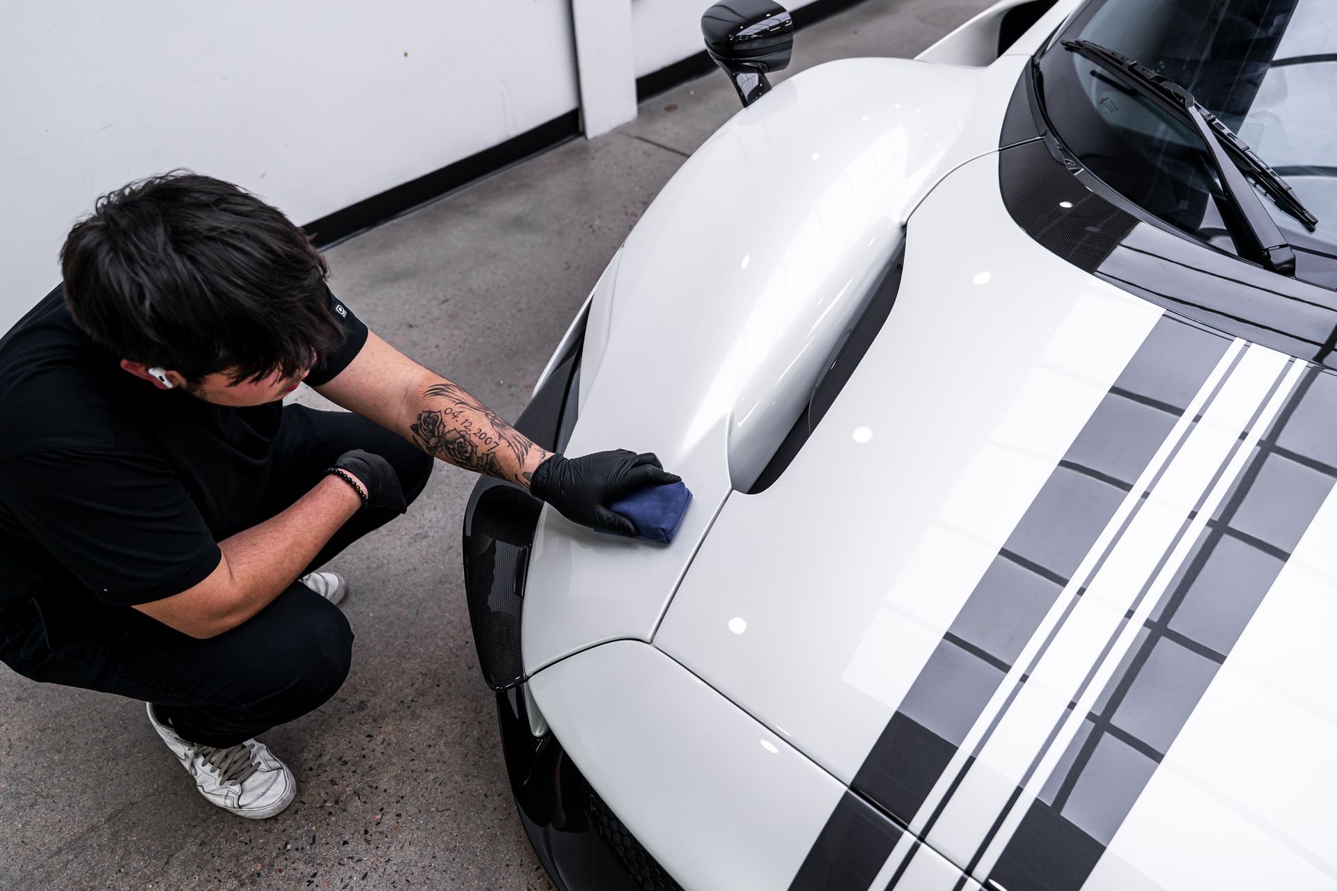 A person applying a coating to a white car's hood, wearing gloves and black clothing.