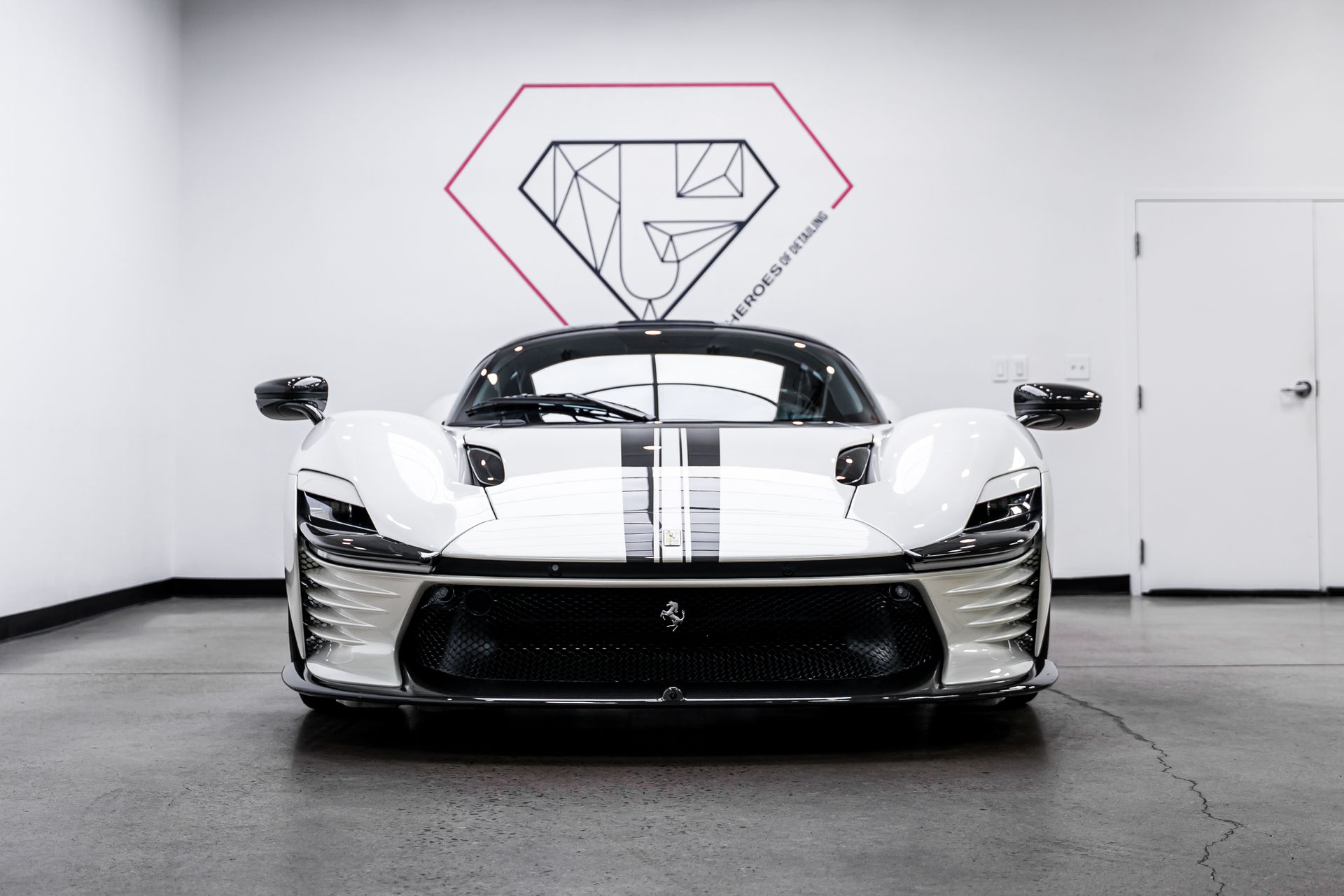 White Ferrari sports car with black racing stripes parked inside a garage, in front of a diamond logo.
