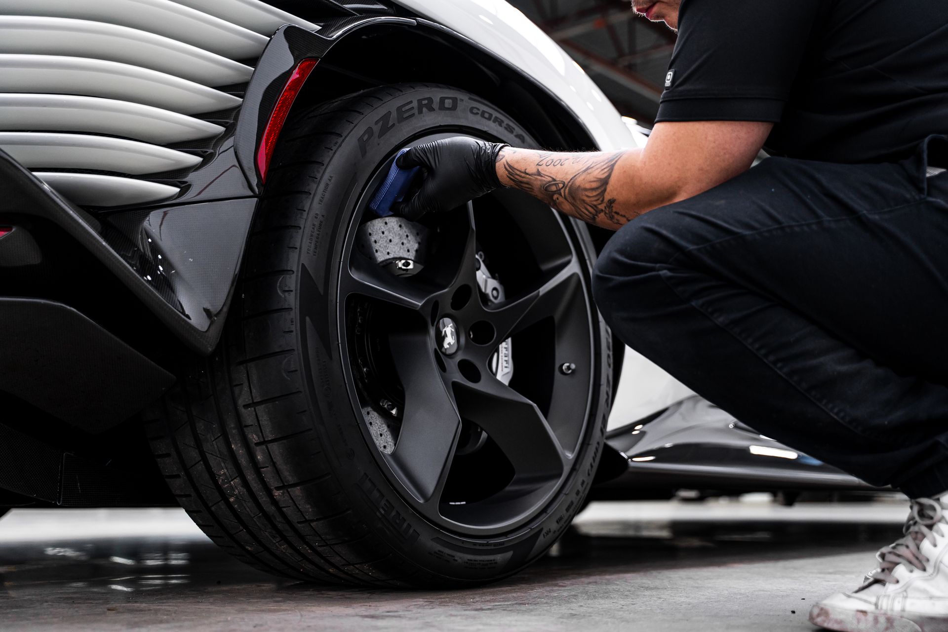 Person cleaning a car's tire with a sponge. Black gloves, black wheel, and a black and white car in a garage.