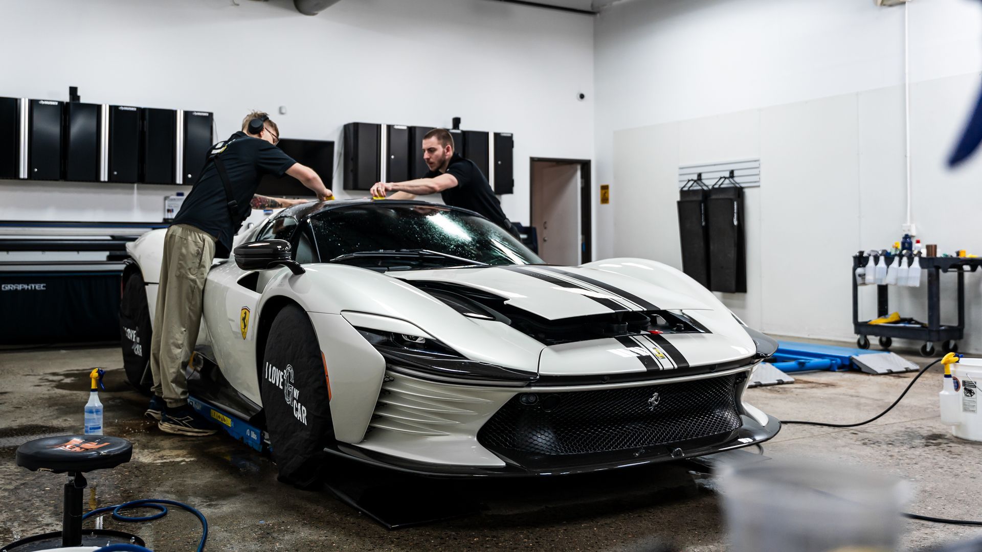 Two men working on a white Ferrari in a garage; applying a protective film.