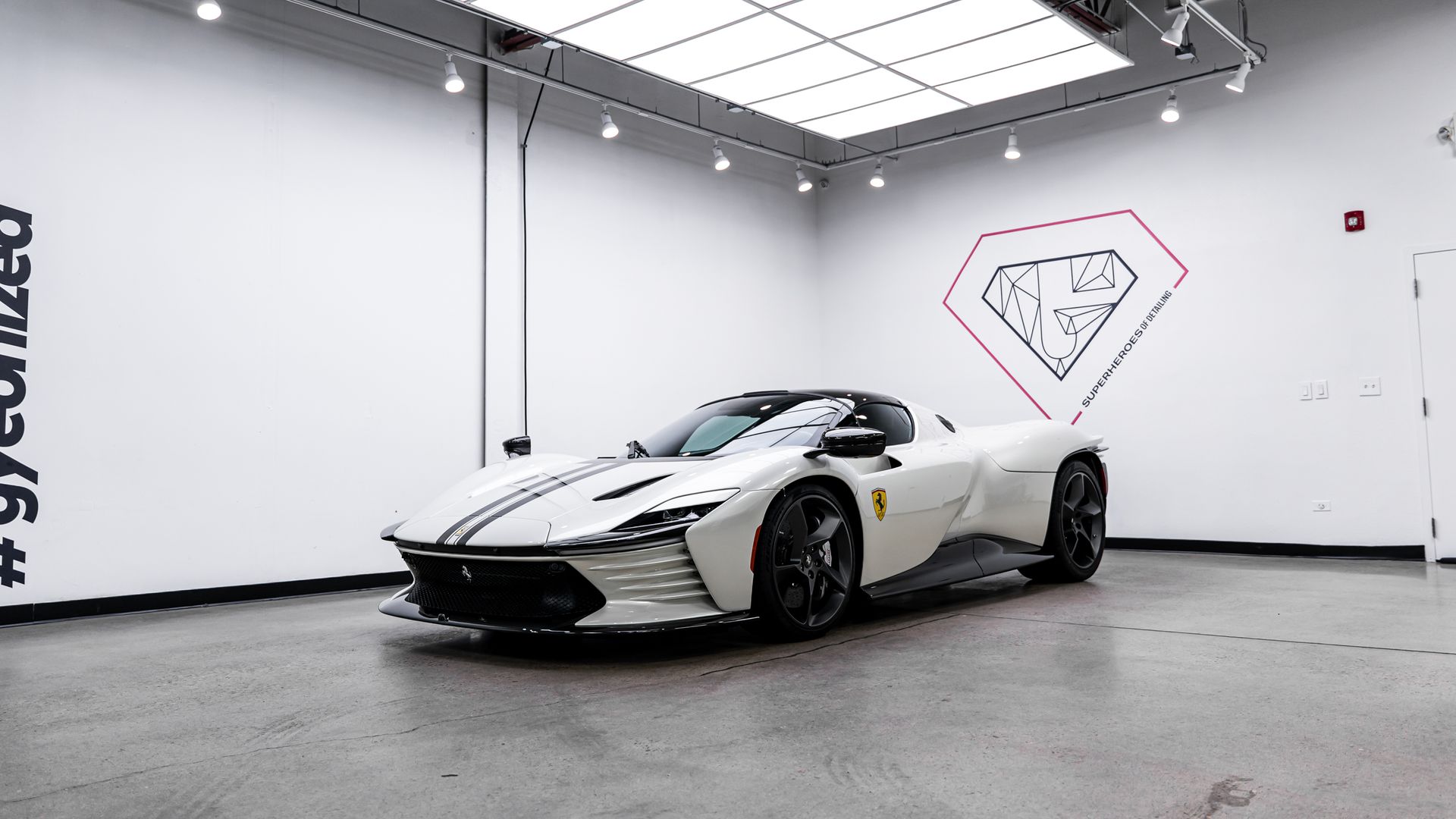 White Ferrari sports car parked inside a white showroom. Black stripes and wheels. Diamond logo on the wall.