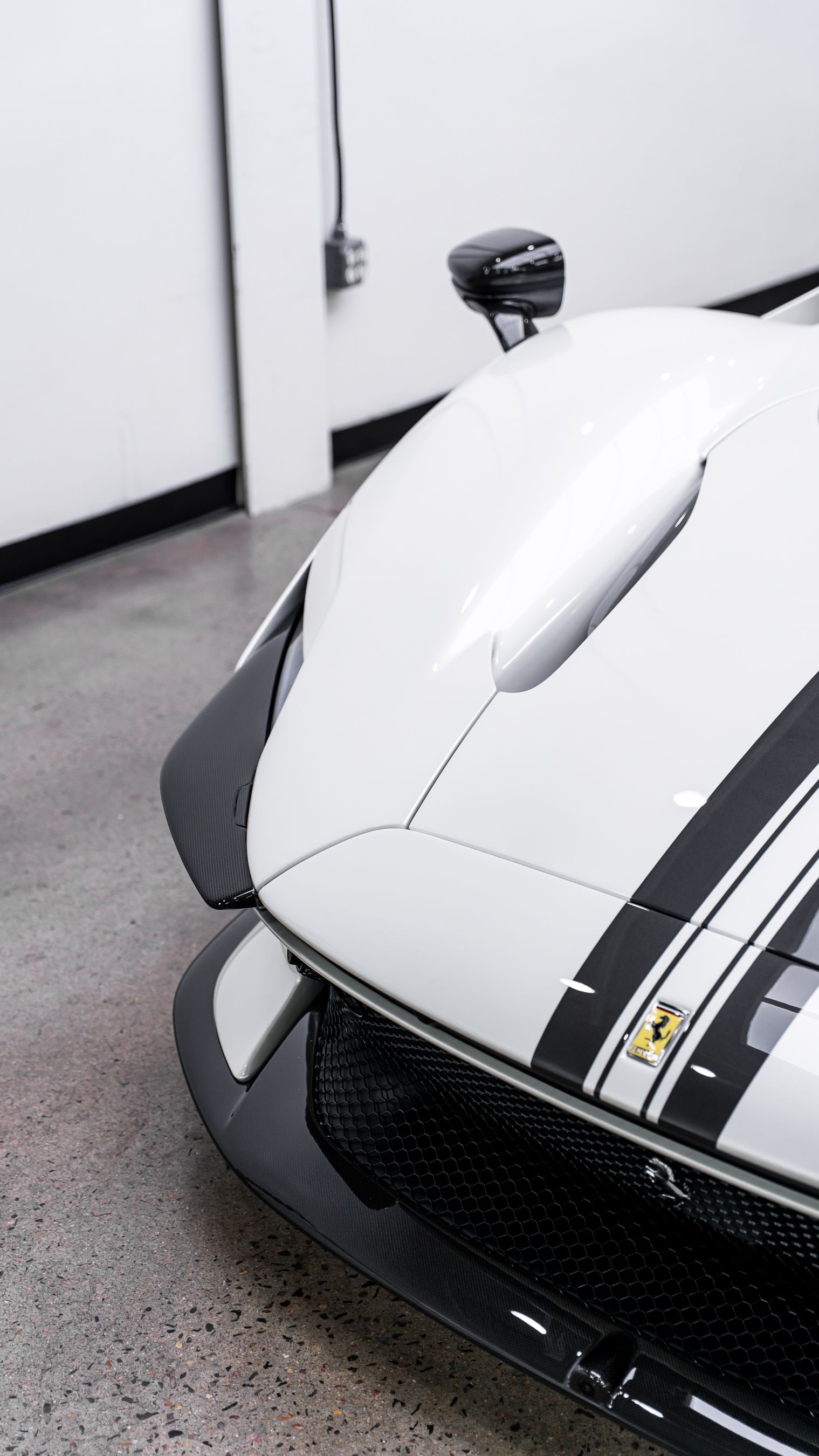 White Ferrari sports car with black racing stripes, angled front view, in a garage.