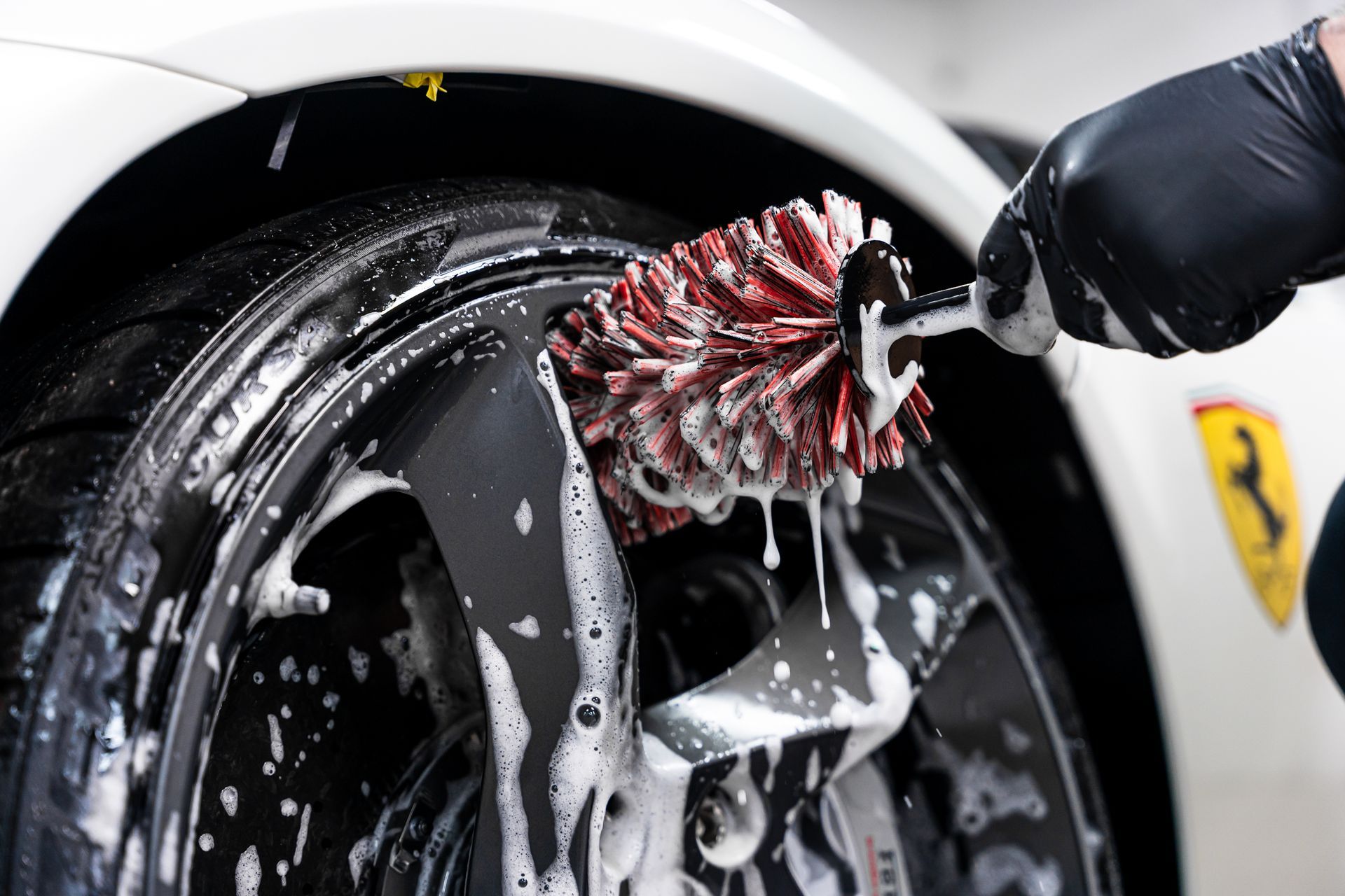 Washing a white car's black rim with a brush, soapy foam visible. Person wearing black glove, near Ferrari logo.