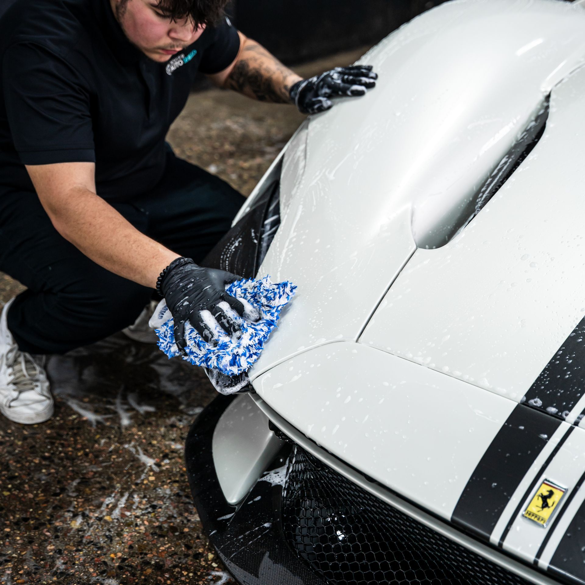 Man washing a white Ferrari with a blue cloth; black gloves on, outdoors.