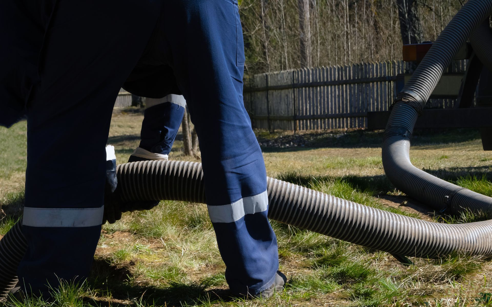 Workers in blue pants handling large black hoses on grass near a fence Workers in blue pants handling large black hoses on grass near a fence