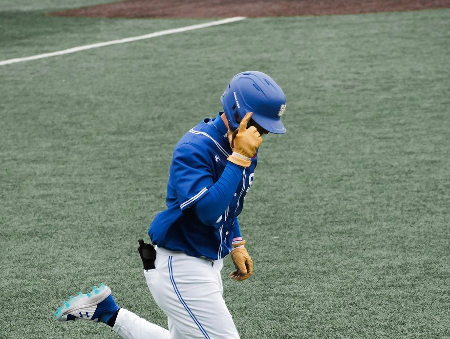 Baseball player in blue uniform kneeling on a field, holding a hand to his helmet.