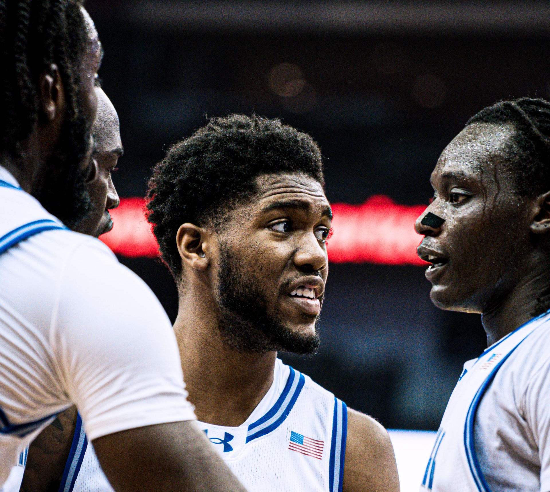Three basketball players in white jerseys huddle and talk on the court
