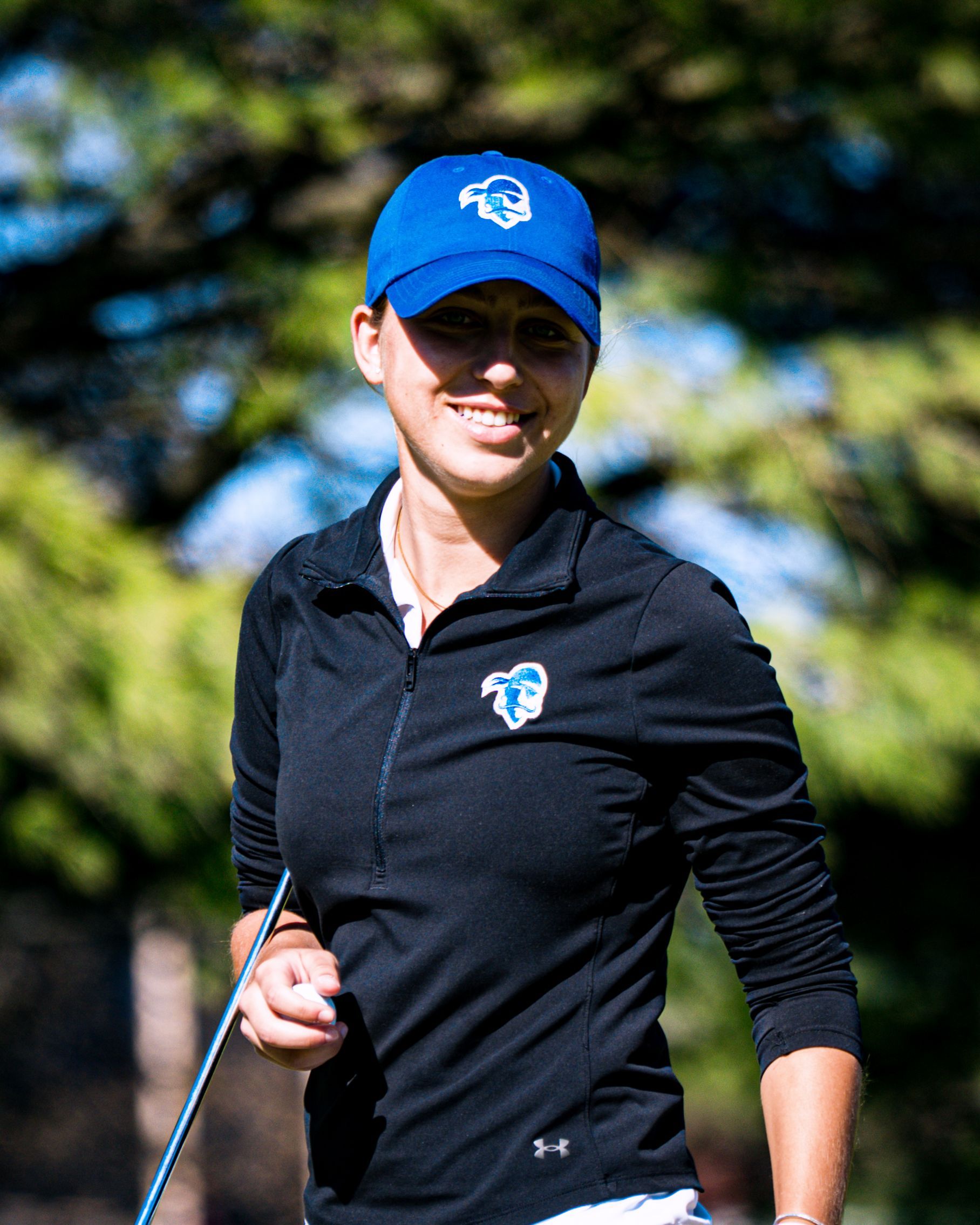 Smiling golfer in blue cap and black pullover, holding a golf club outdoors on a sunny course