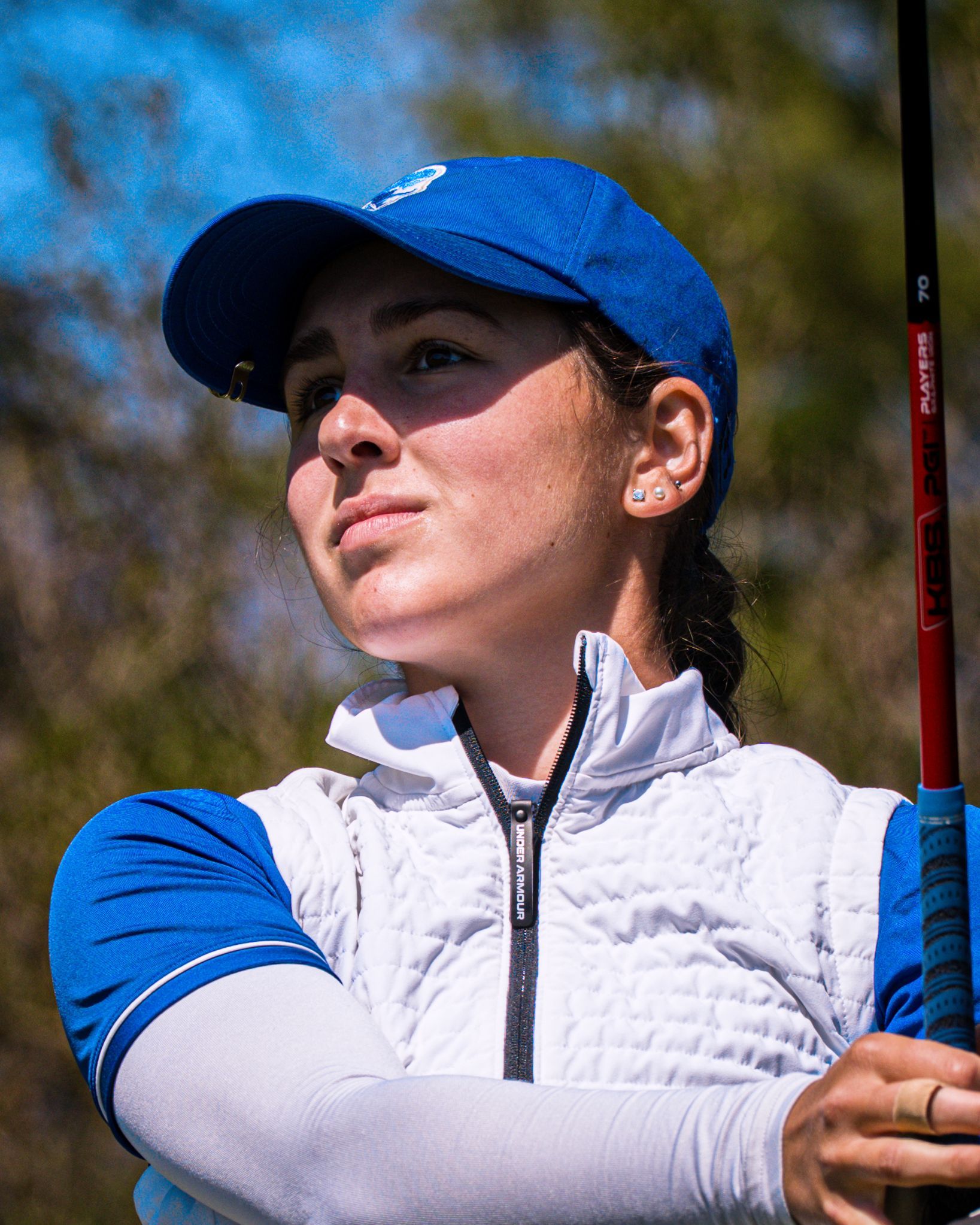 Golfer in blue cap and jacket holding a club, looking to the side outdoors