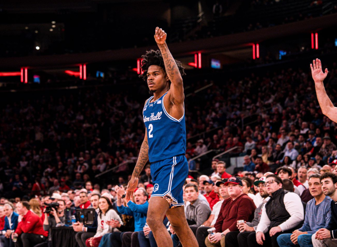 Basketball player in blue jersey raises a hand near the court sidelines in a packed arena