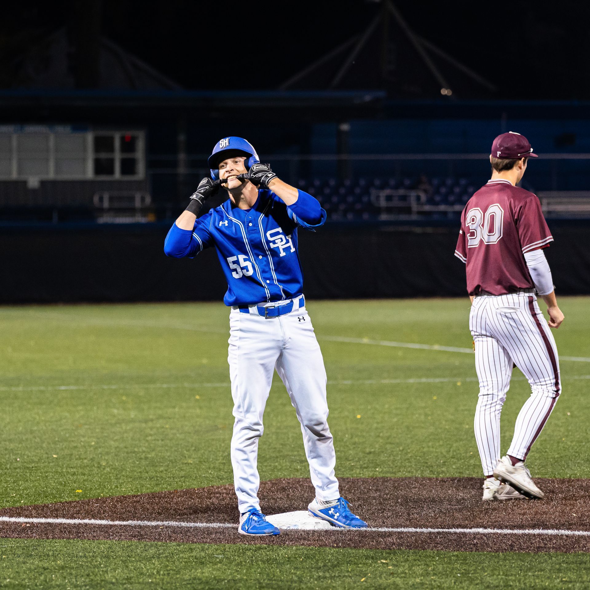 Two baseball players on the field at night, one in blue adjusting his glove and one in maroon standing nearby