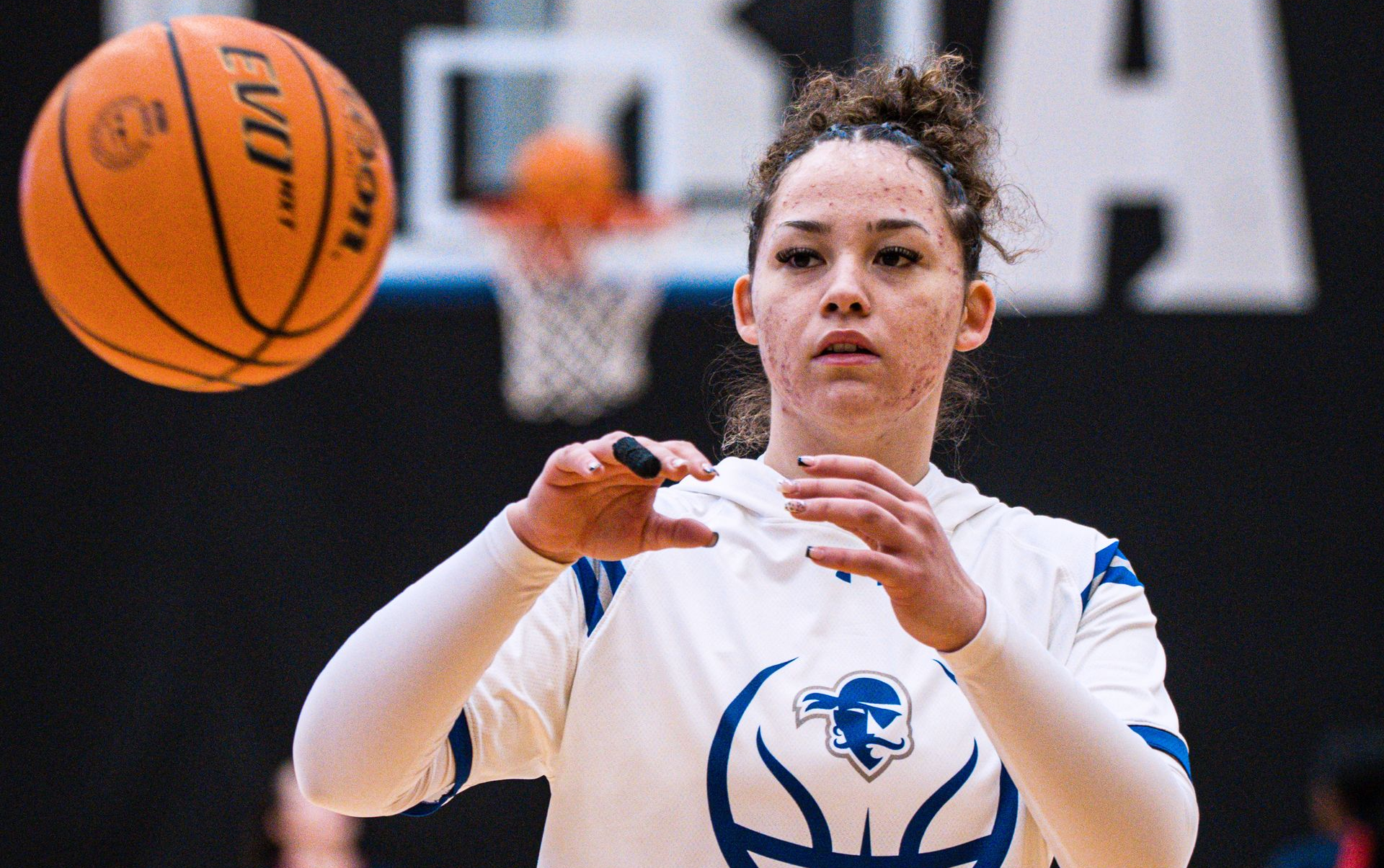Basketball player in white uniform catches a ball during practice, with hoop blurred in the background.