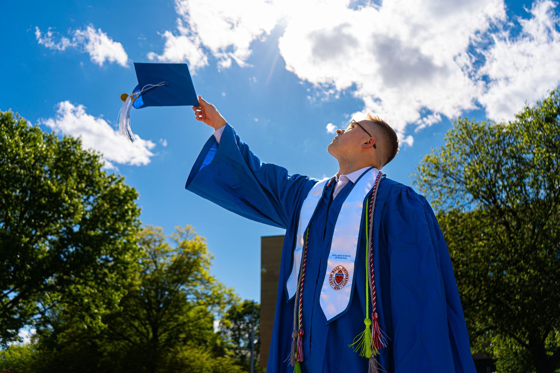 Graduate in blue gown tossing cap into the sky on a sunny outdoor campus lawn