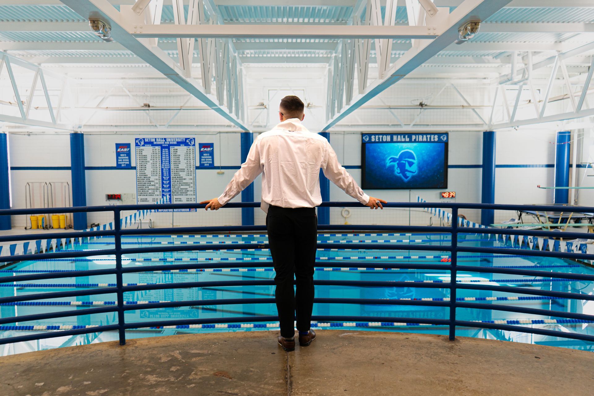 Man standing on an indoor pool balcony, looking over blue lanes and bright white beams