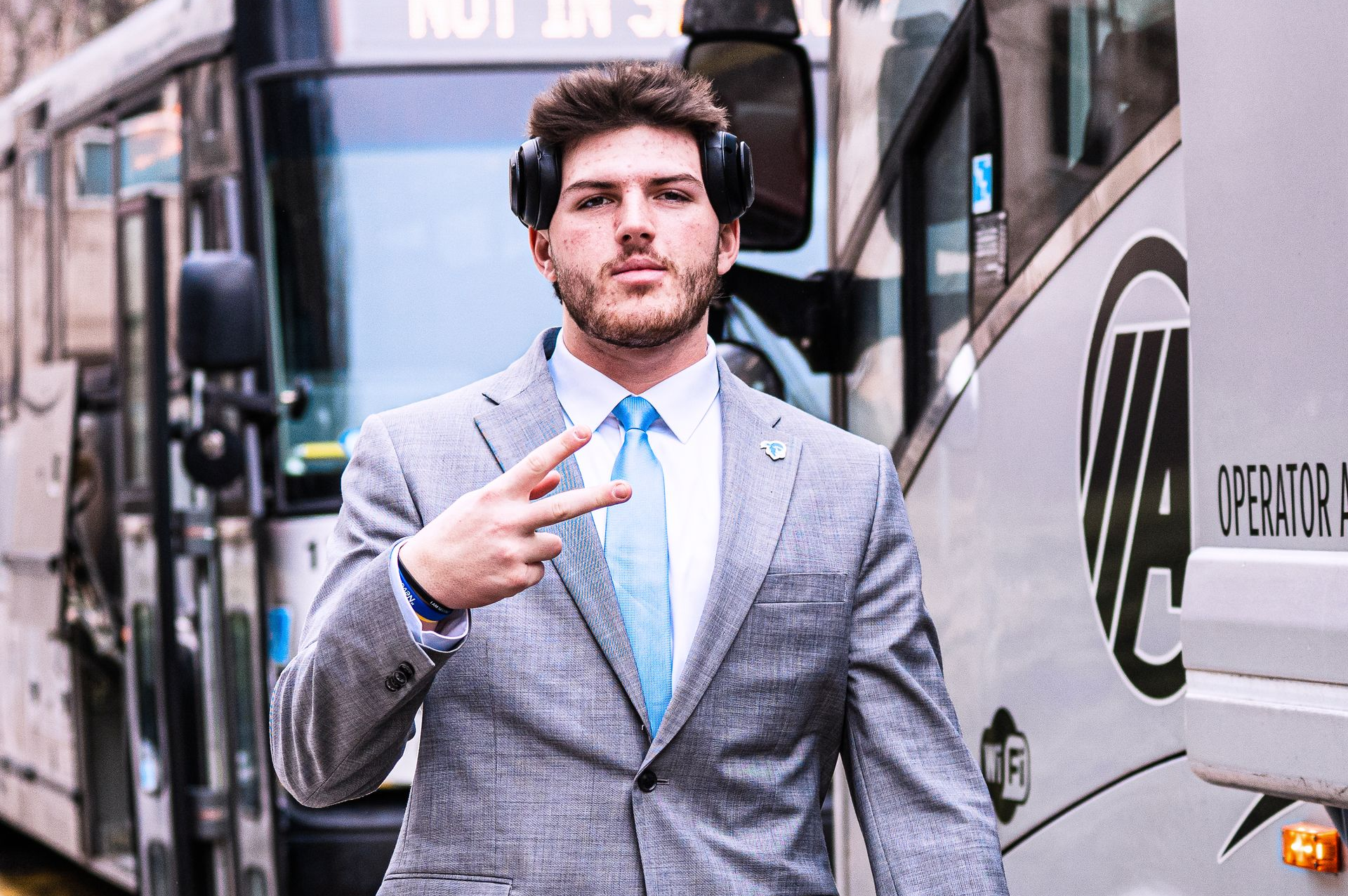 Man in a gray suit making a peace sign on a city street beside a bus