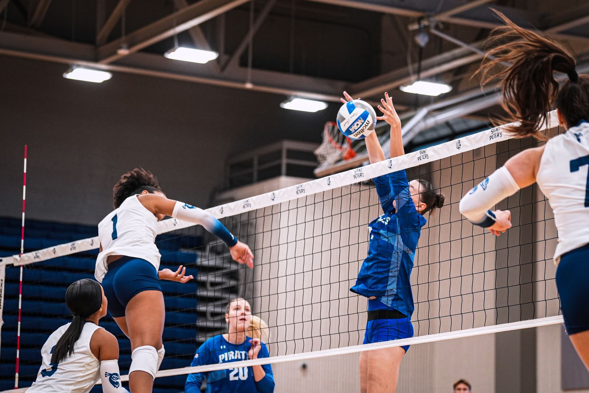 Volleyball players at the net during an indoor match, with a blue-clad blocker reaching to defend a spike