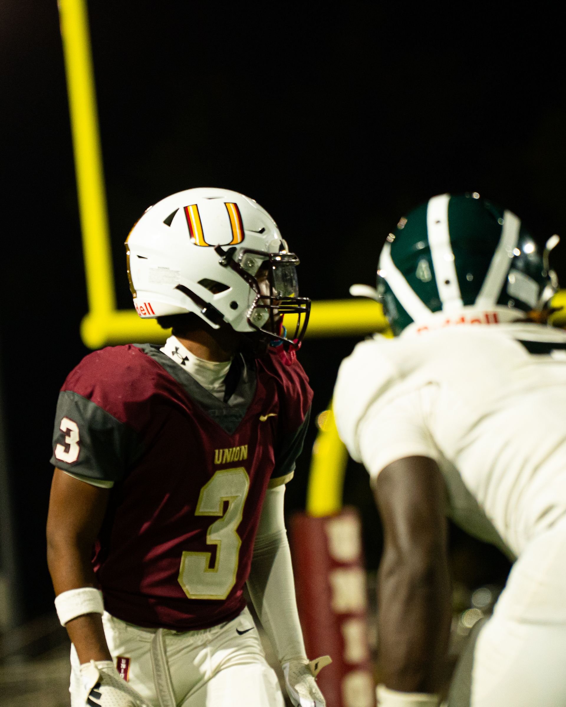 Football players in maroon and white uniforms face off under stadium lights at night.