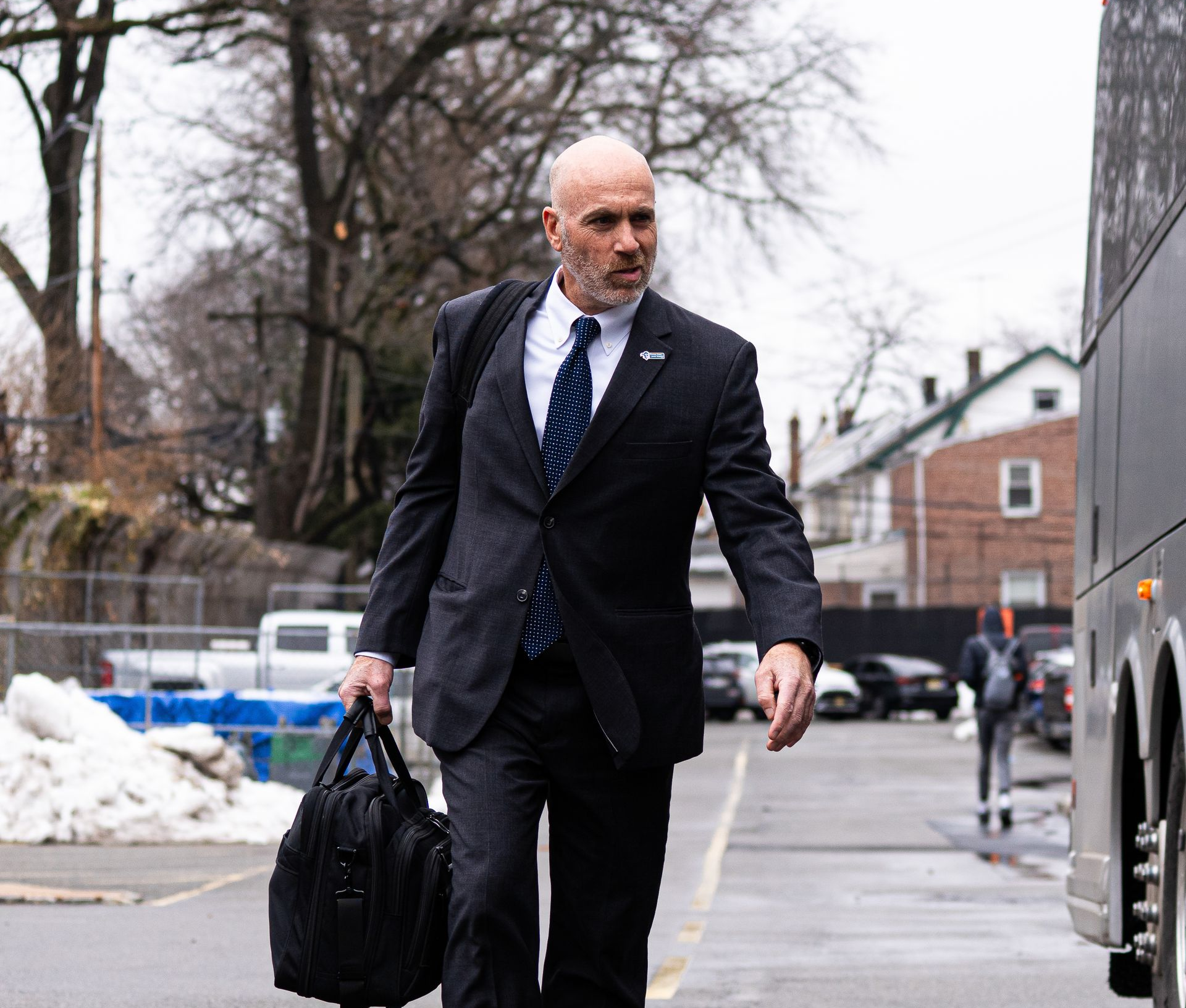Man in dark suit carrying a black bag on a city sidewalk beside a bus, with snow and bare trees nearby