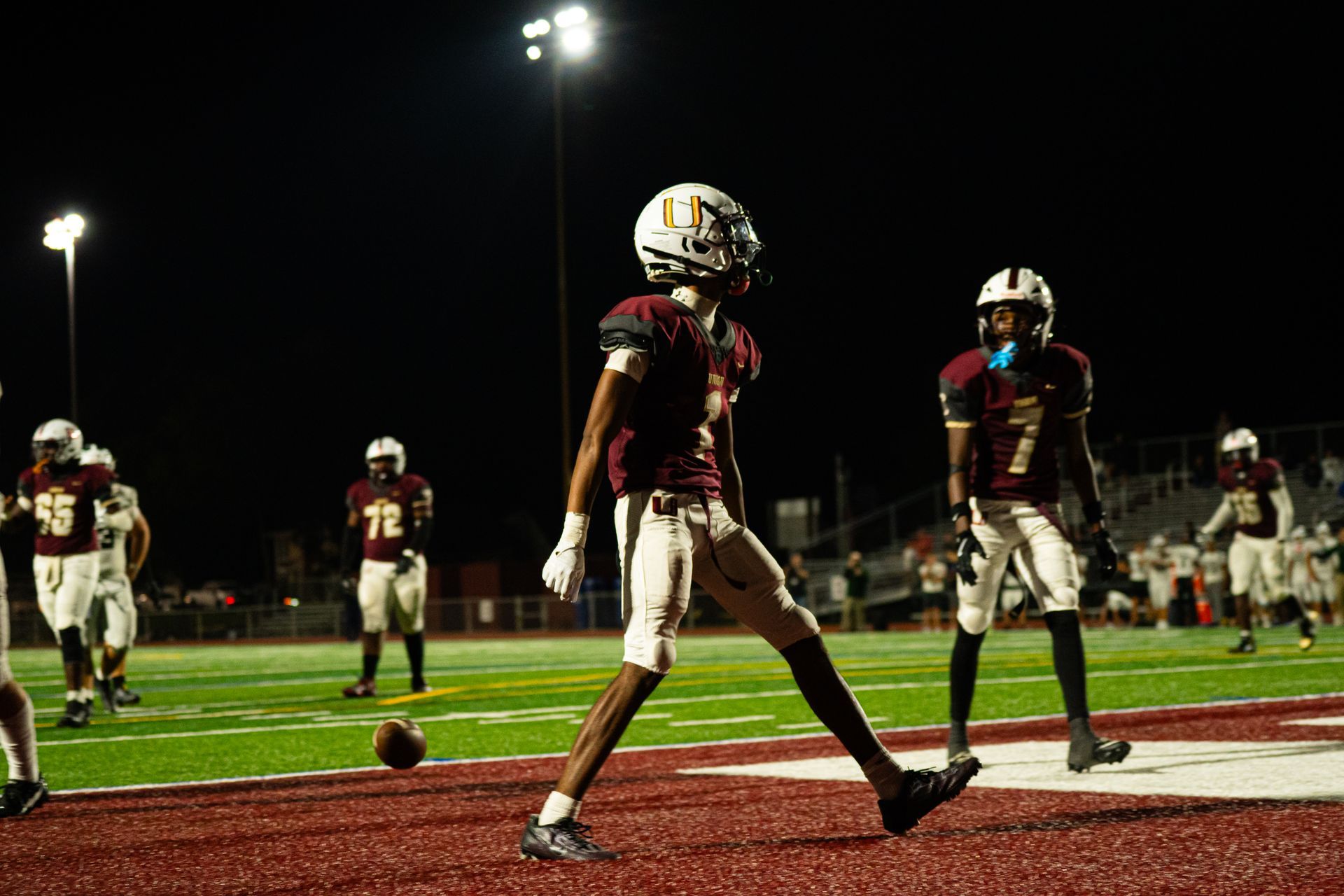 Football players on a lit field at night, with one player walking near the goal line.