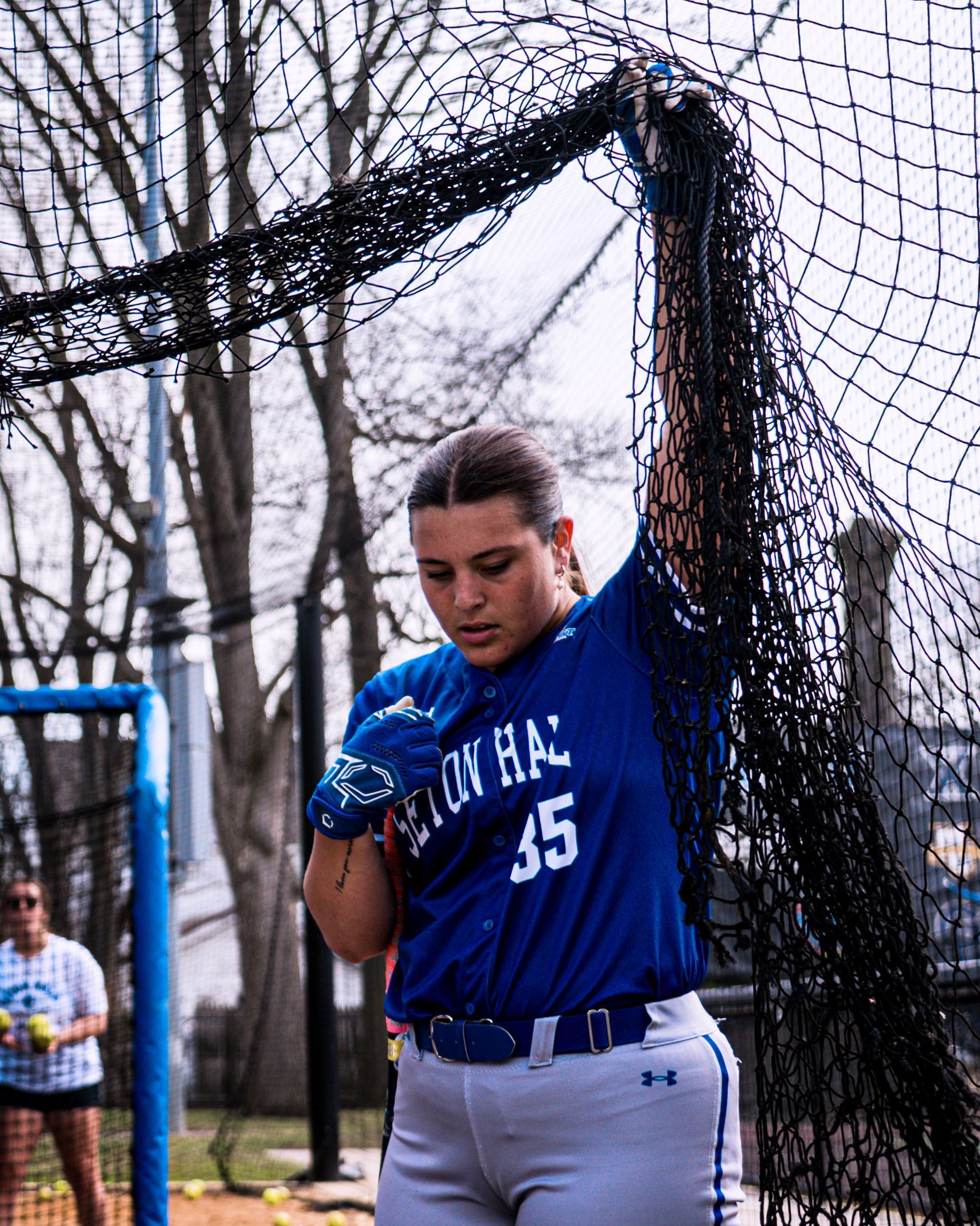 Softball player in blue jersey and gloves stands in a batting cage holding up the netting.