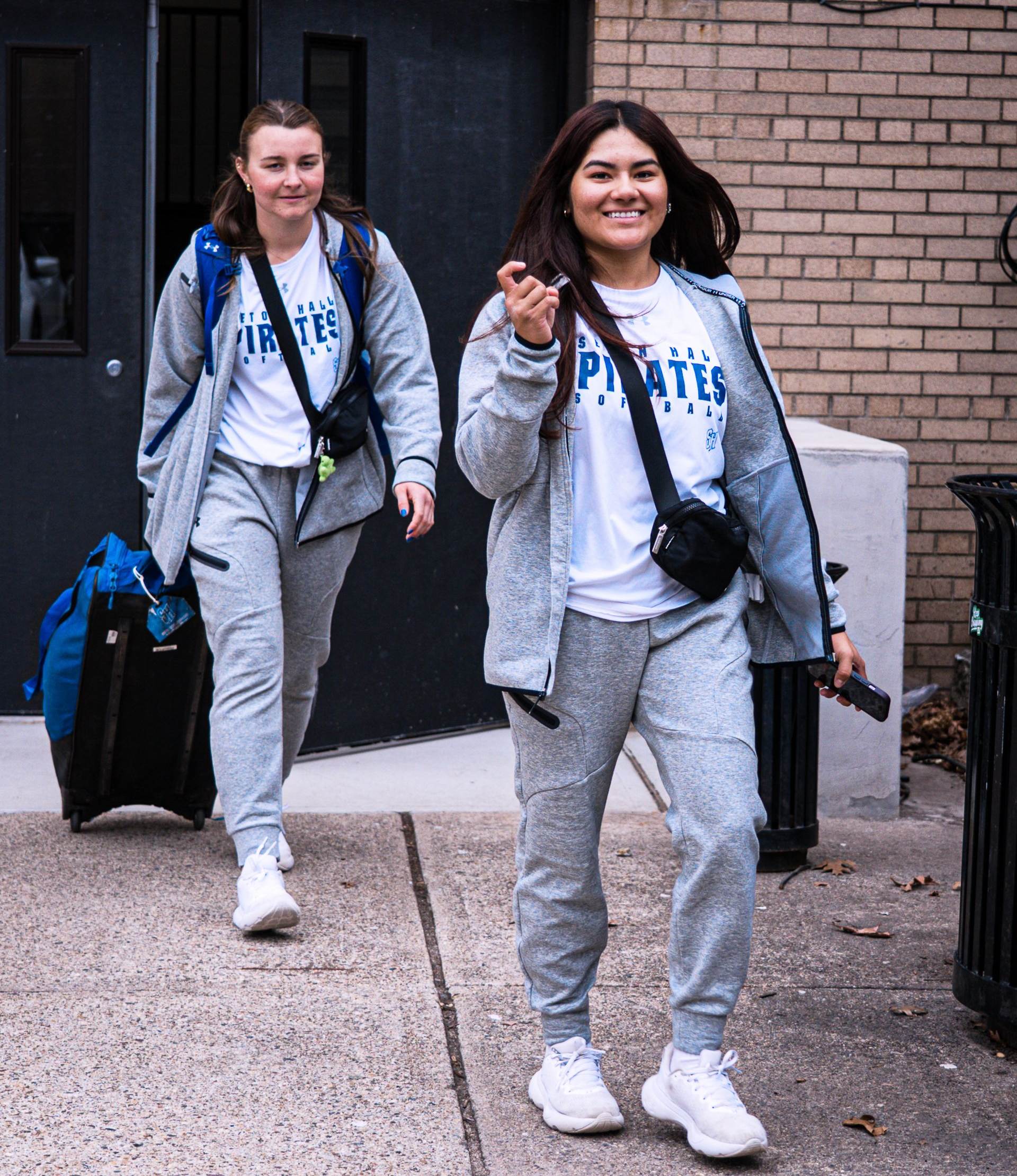 Two women in matching gray tracksuits walk outdoors with luggage, smiling and carrying bags.