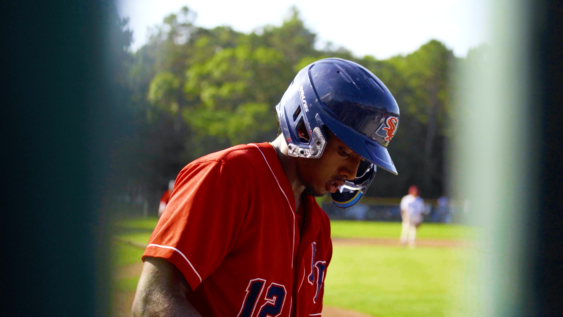 Baseball player in red jersey and blue helmet looking down on a sunny field