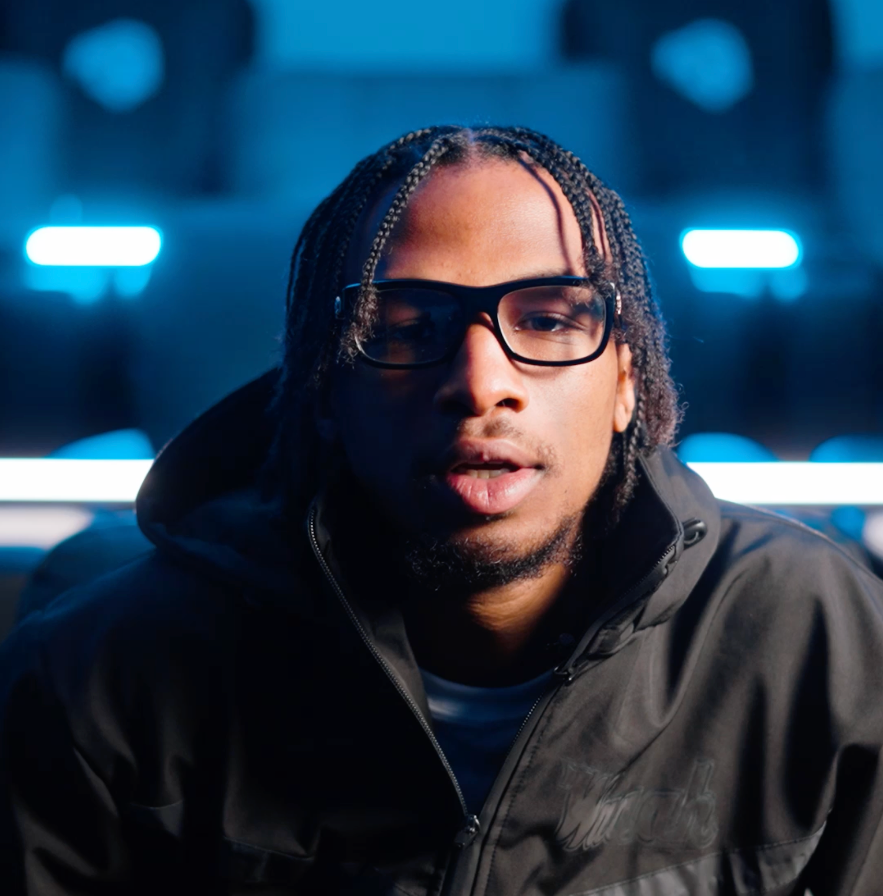 Person with braided hair and glasses in a dark jacket, lit by blue neon lights.