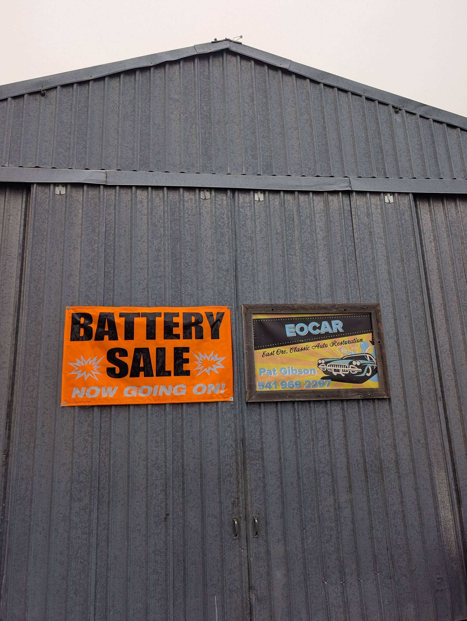 Gray metal building with orange “Battery Sale” sign and a small racing poster on the wall