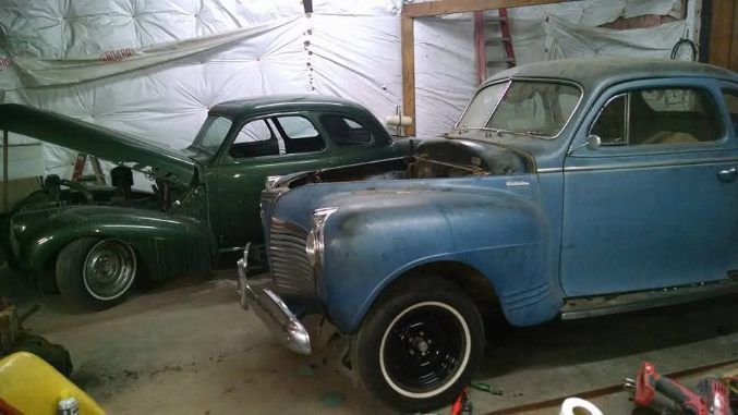 Two vintage cars, one blue and one dark green, parked inside a garage or barn.