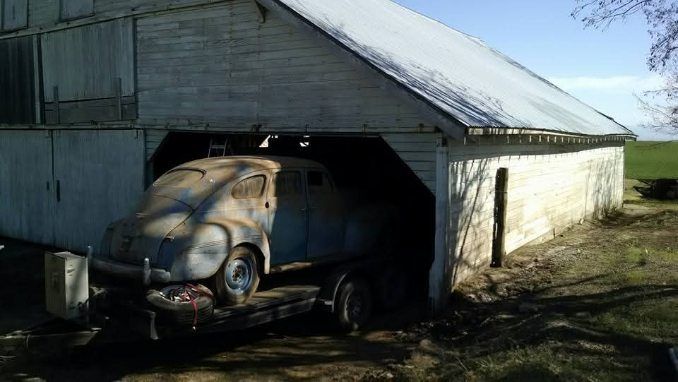 Old rusted car parked inside a weathered wooden shed with an open doorway and bright sunlight outside