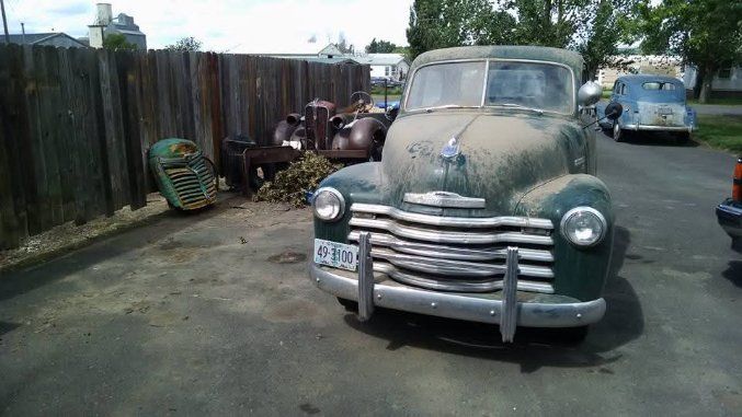Dusty vintage green car parked on a street beside a fence, with another blue car in the background