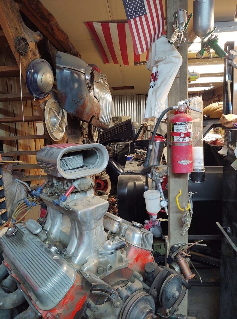 Vintage car engine and tools in a cluttered workshop with an American flag hanging above