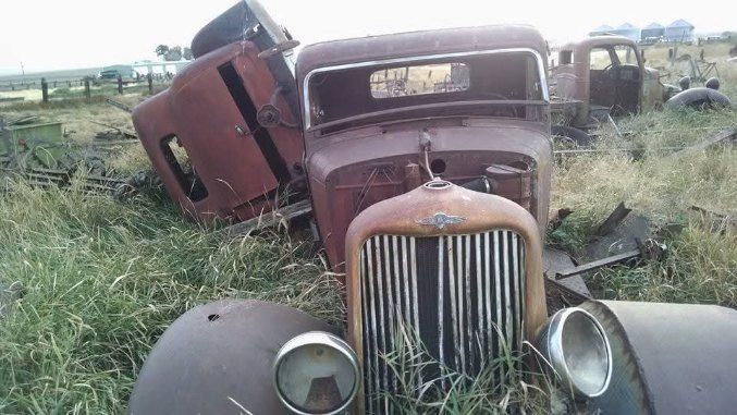 Rusty vintage truck wrecks in a grassy field, front grille facing forward with another damaged vehicle behind.