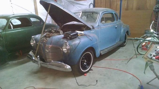 Blue vintage car in a garage with its hood open, parked beside a green car and a bicycle.