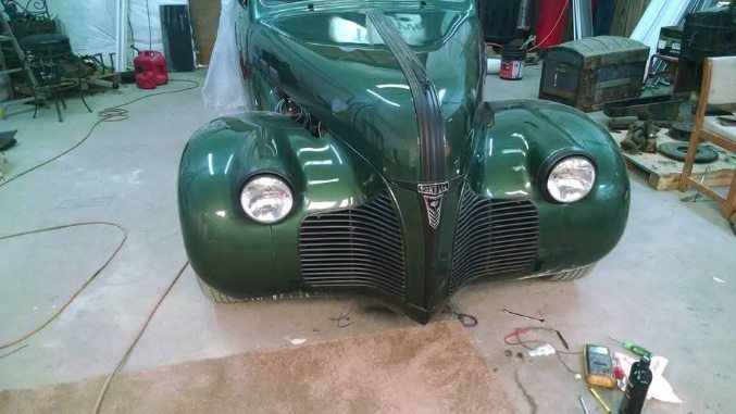 Front view of a dark green vintage car in a workshop, with round headlights and exposed grille.