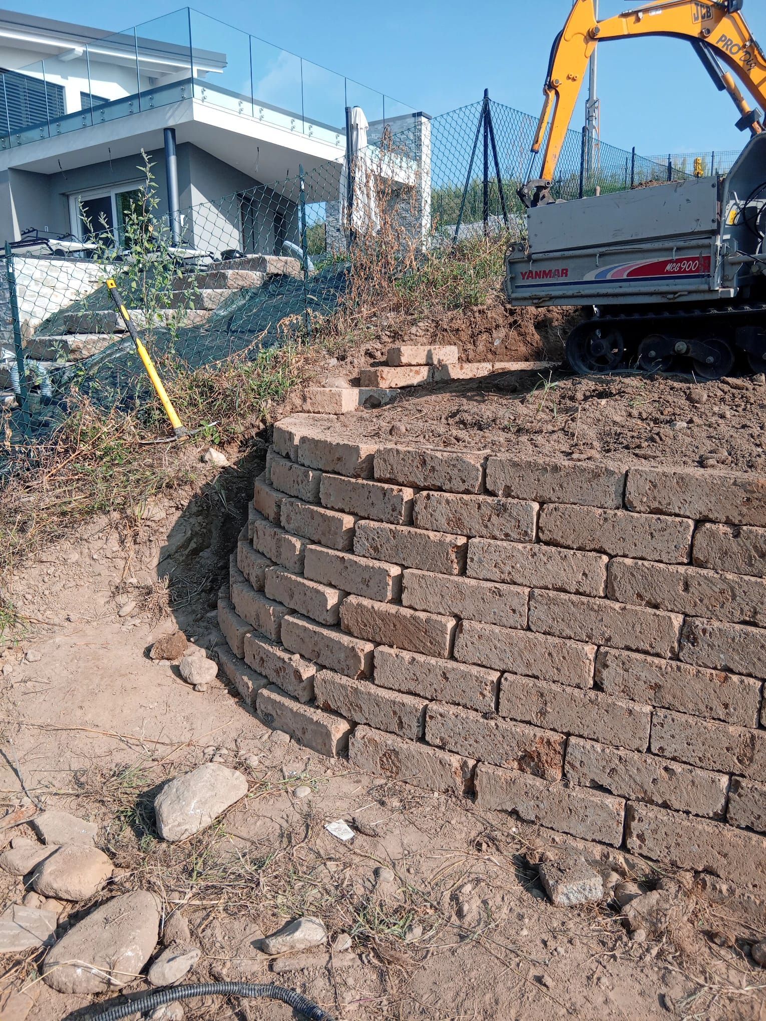 Muro di contenimento curvo in pietra a secco accanto a una casa, con un camion e un escavatore su un pendio di terra.