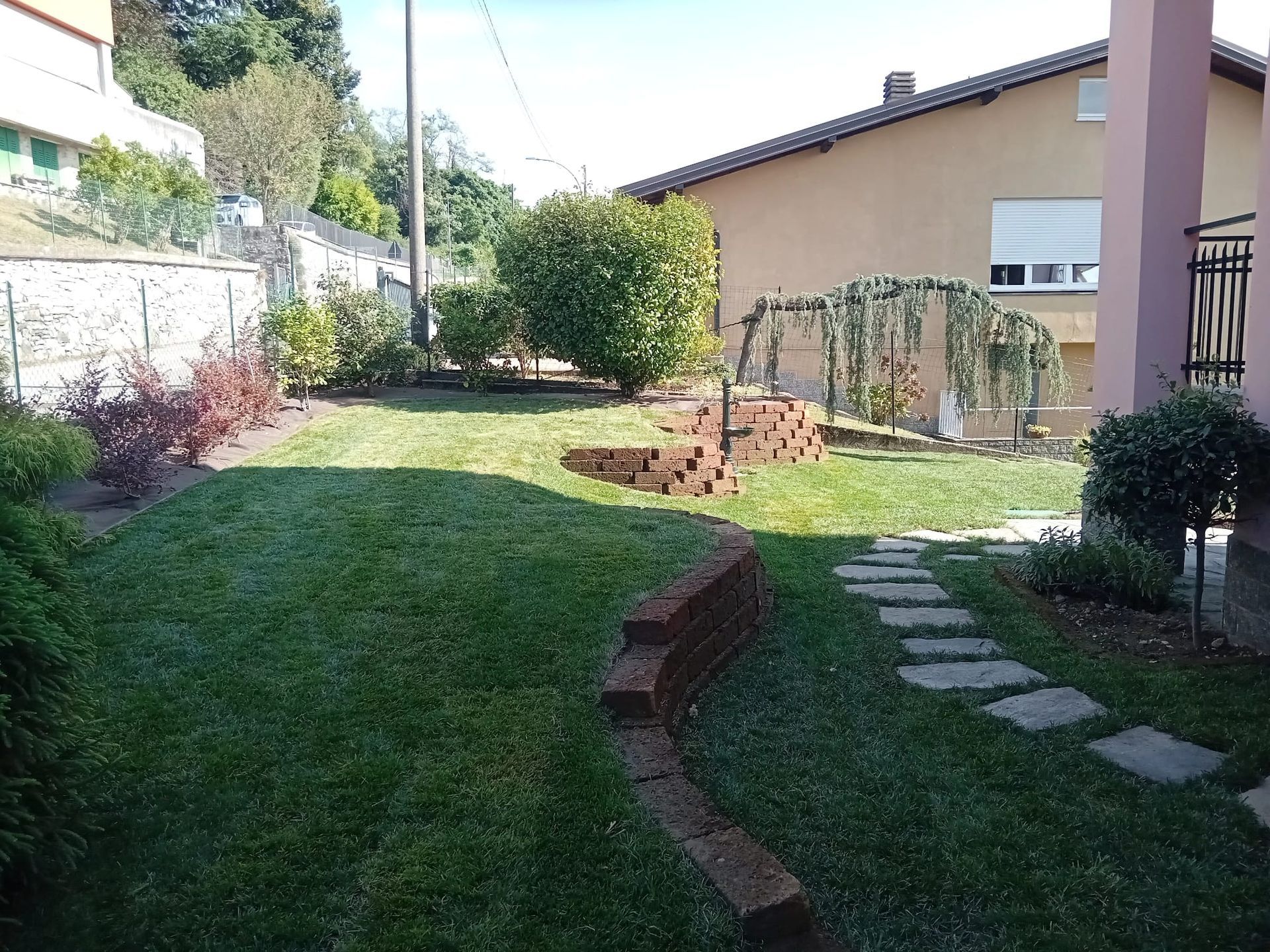 Giardino sul retro con prato, sentiero in pietra, bordure in mattoni, arbusti e patio di una casa sullo sfondo.