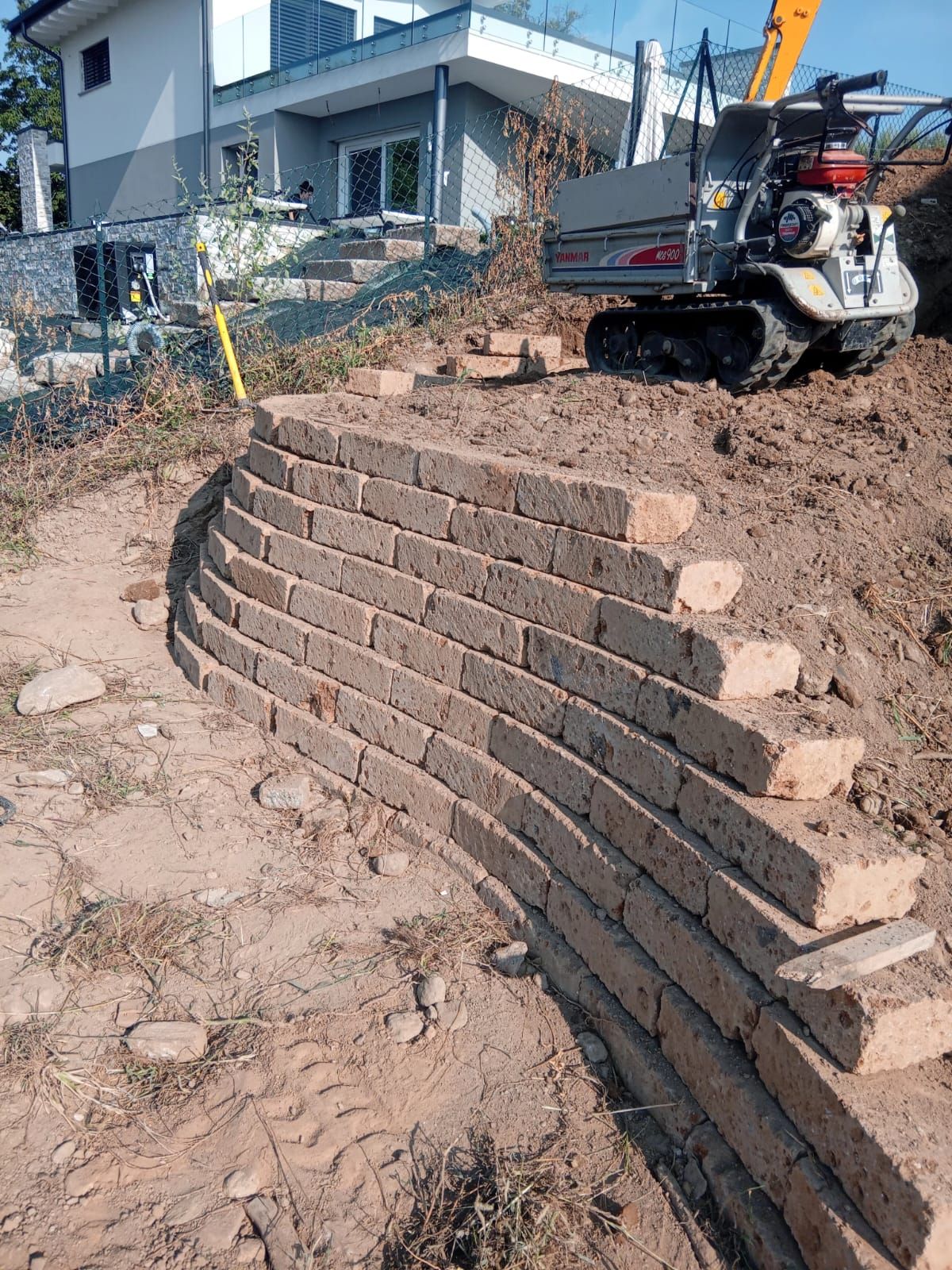 Cantiere edile con un muro di contenimento curvo in mattoni in costruzione accanto a una casa e un escavatore.