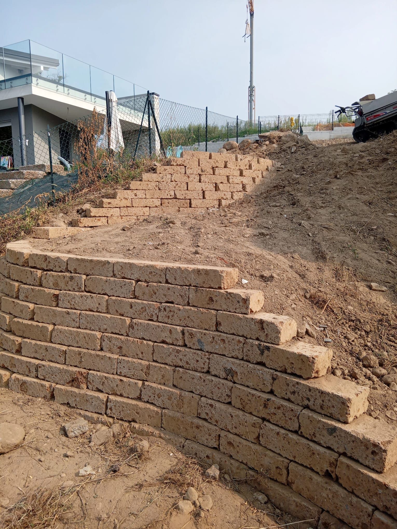 Muro di contenimento in pietra in costruzione su un pendio di terra accanto a un edificio e a una recinzione.