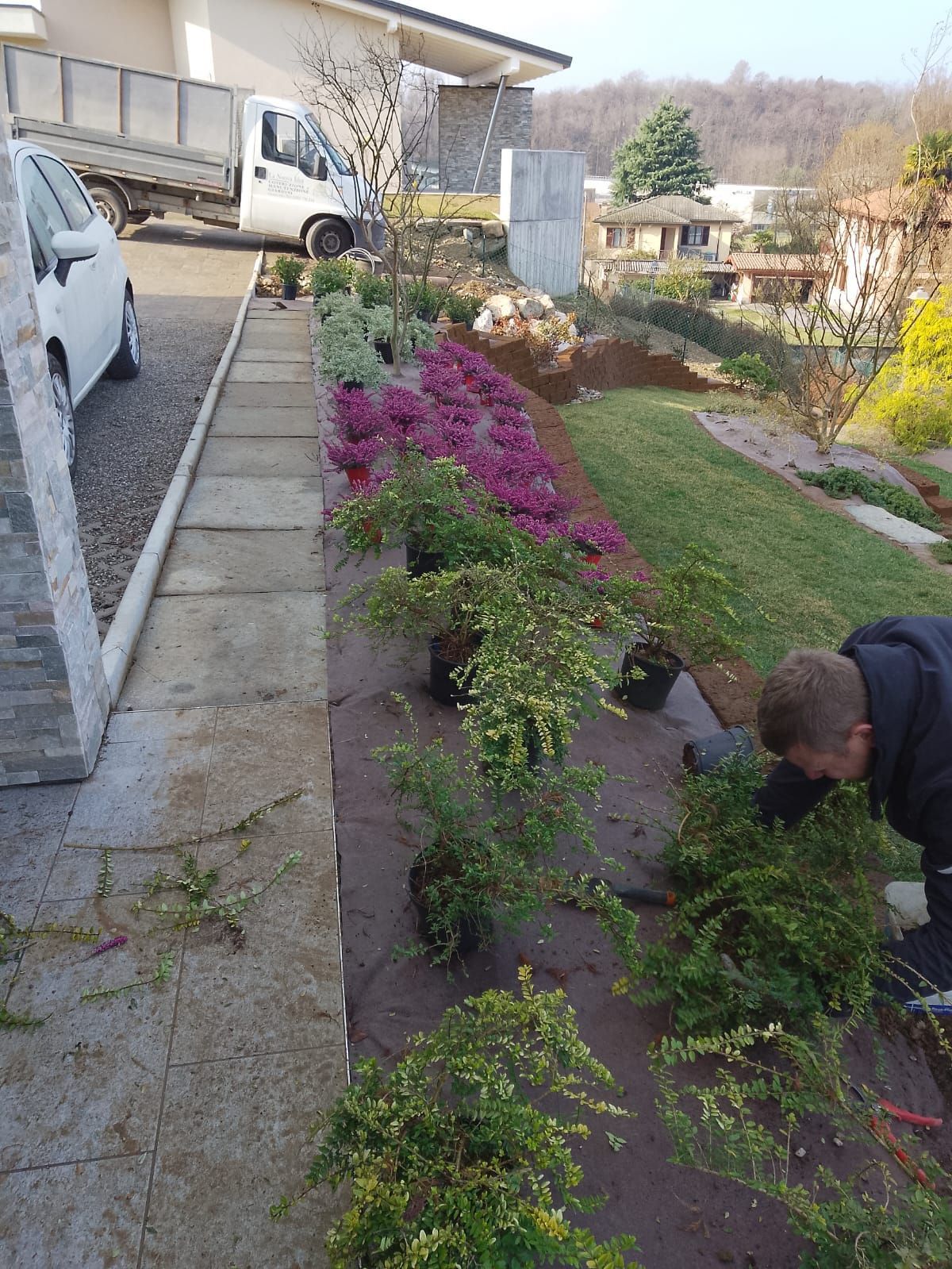 Una persona si dedica al giardinaggio accanto a un sentiero di pietra e a fiori rosa in un cortile in pendenza.