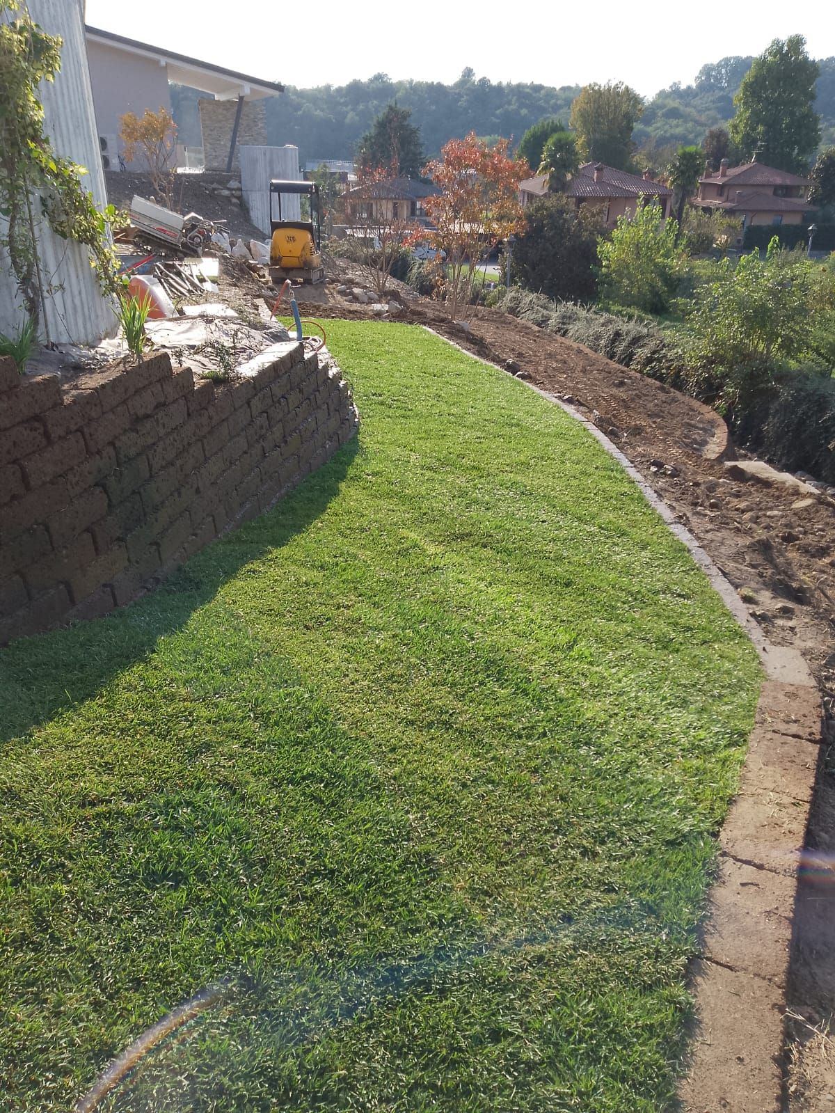 Un prato verde e sinuoso costeggia un muro di pietra e un sentiero da giardino in un cortile soleggiato.