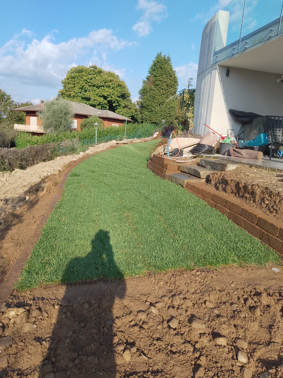 Sentiero erboso in costruzione accanto a un edificio moderno, con bordi di terra e l'ombra di una persona in primo piano.