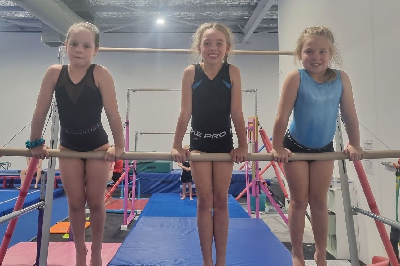 Three young girls are on uneven bars in a gym.