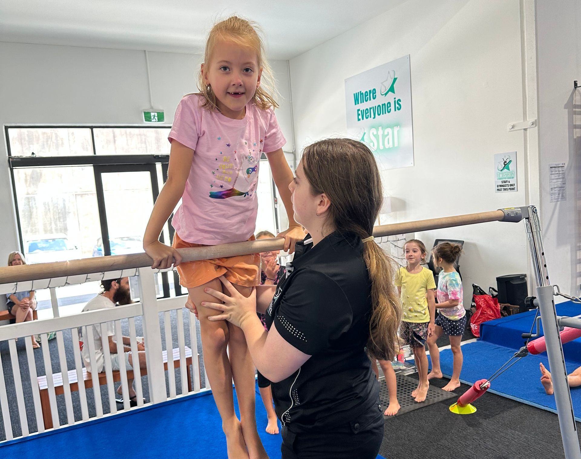 A woman is helping a little girl balance on a bar in a gym.