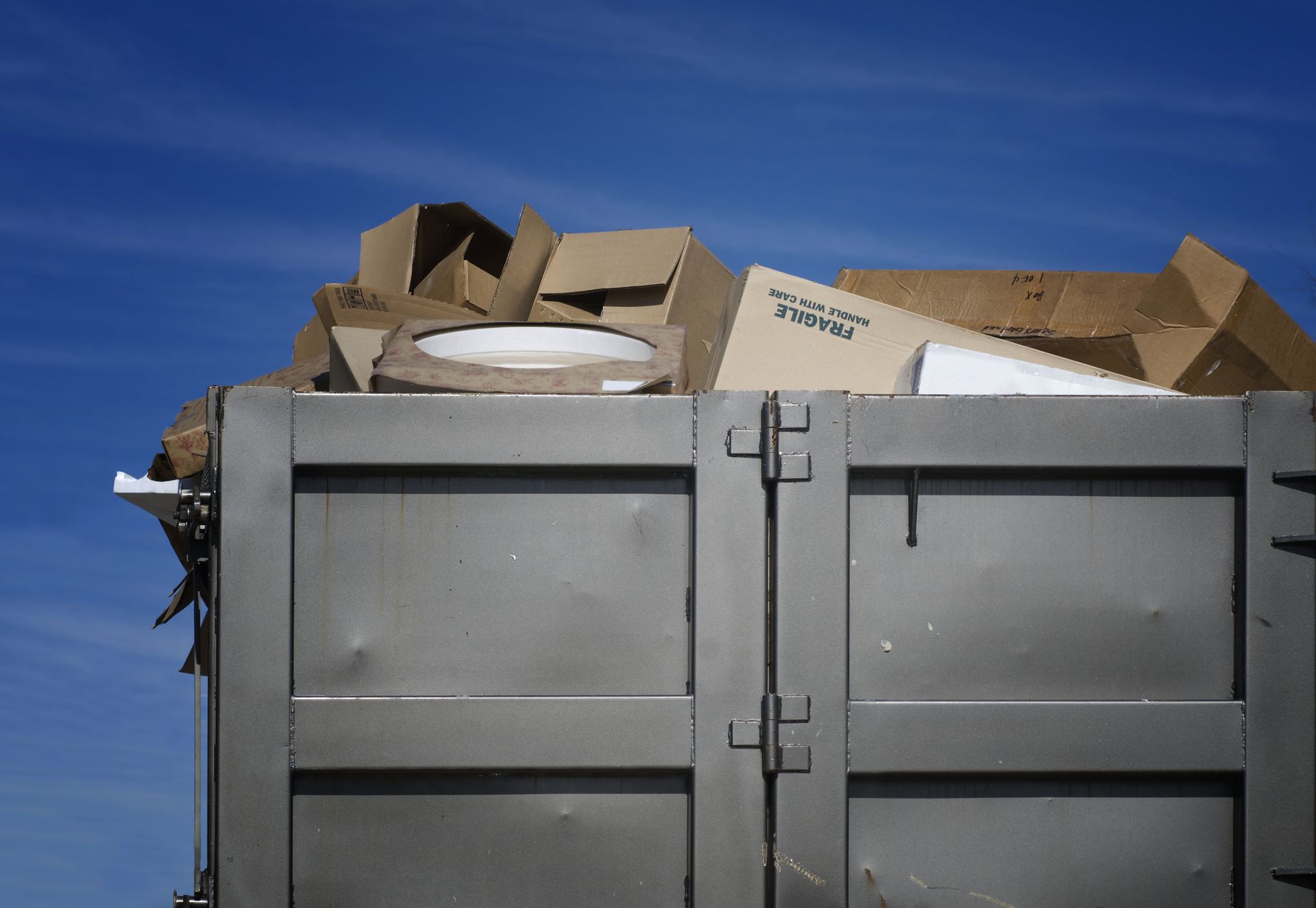 Cardboard boxes overflowing a metal dumpster against a blue sky.