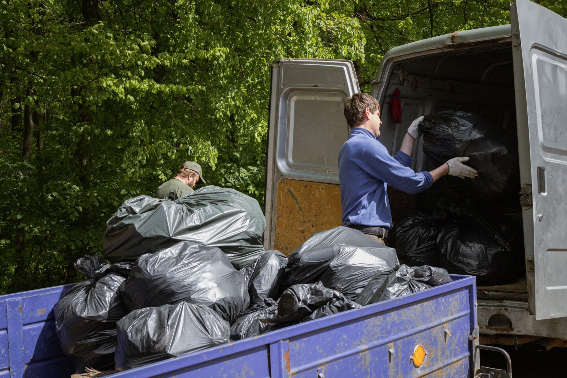 Two people loading black trash bags into a van in a wooded area.